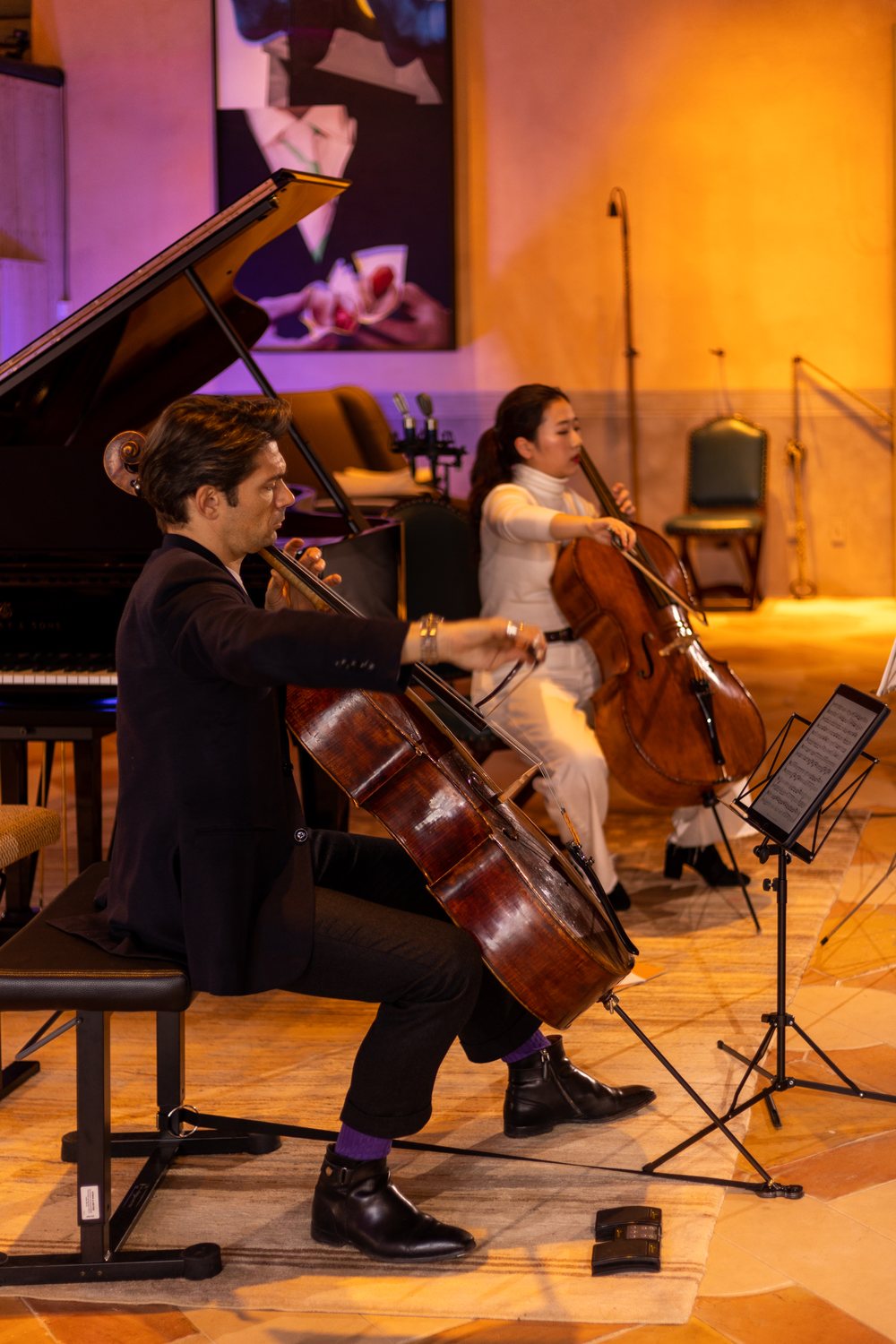 Two cellists playing in the showroom, lit mostly by warm amber light, with a violet glow behind them.