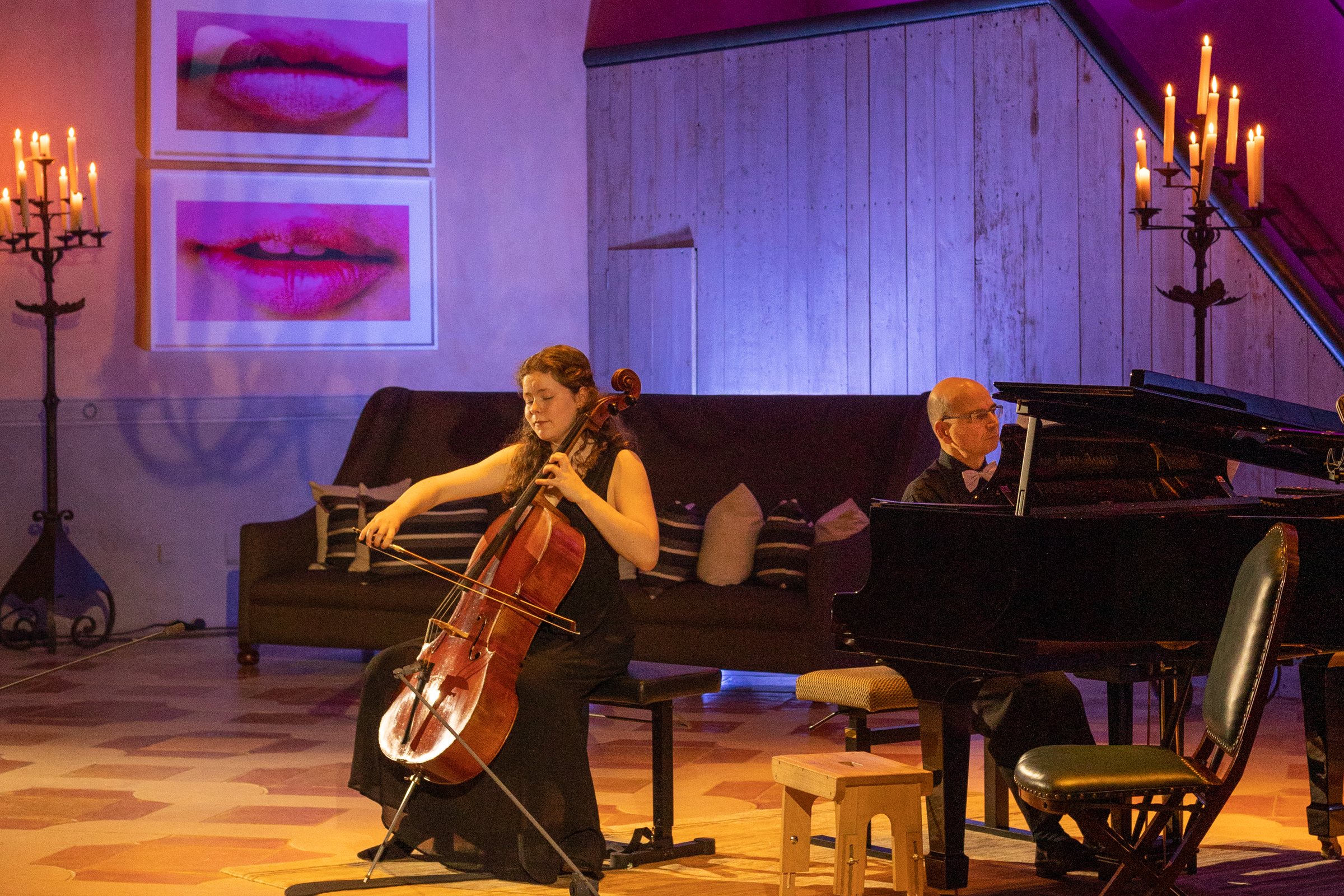 A cellist and a pianist performing in the Classeek showroom. Violet and magenta light washes the wooden walls and two large prints of pink lips; warm amber light falls on the floor; candelabras stand at both sides.